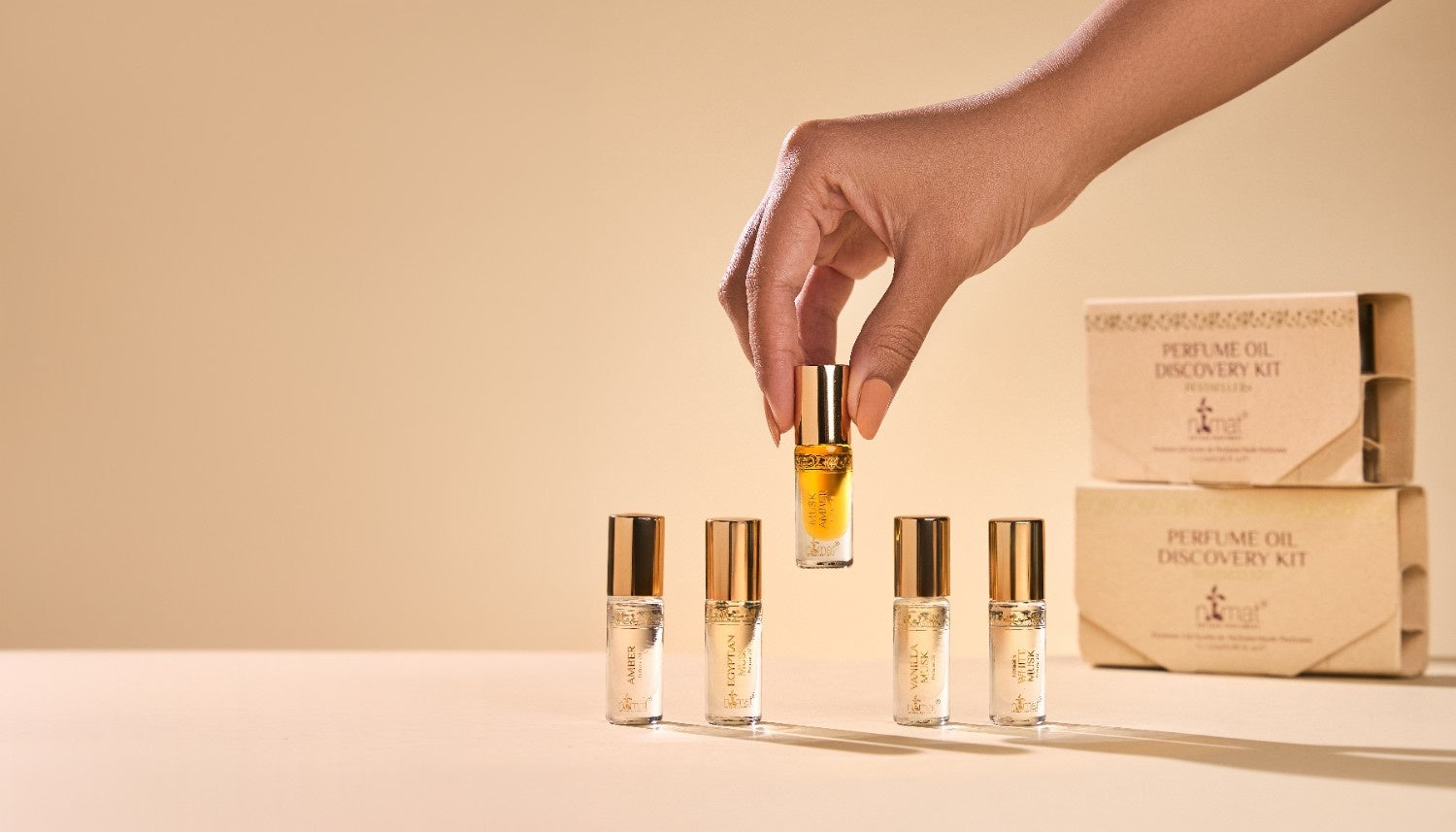 Hand holding a musk amber bottle with a set of other gift bottles and a box on a beige background