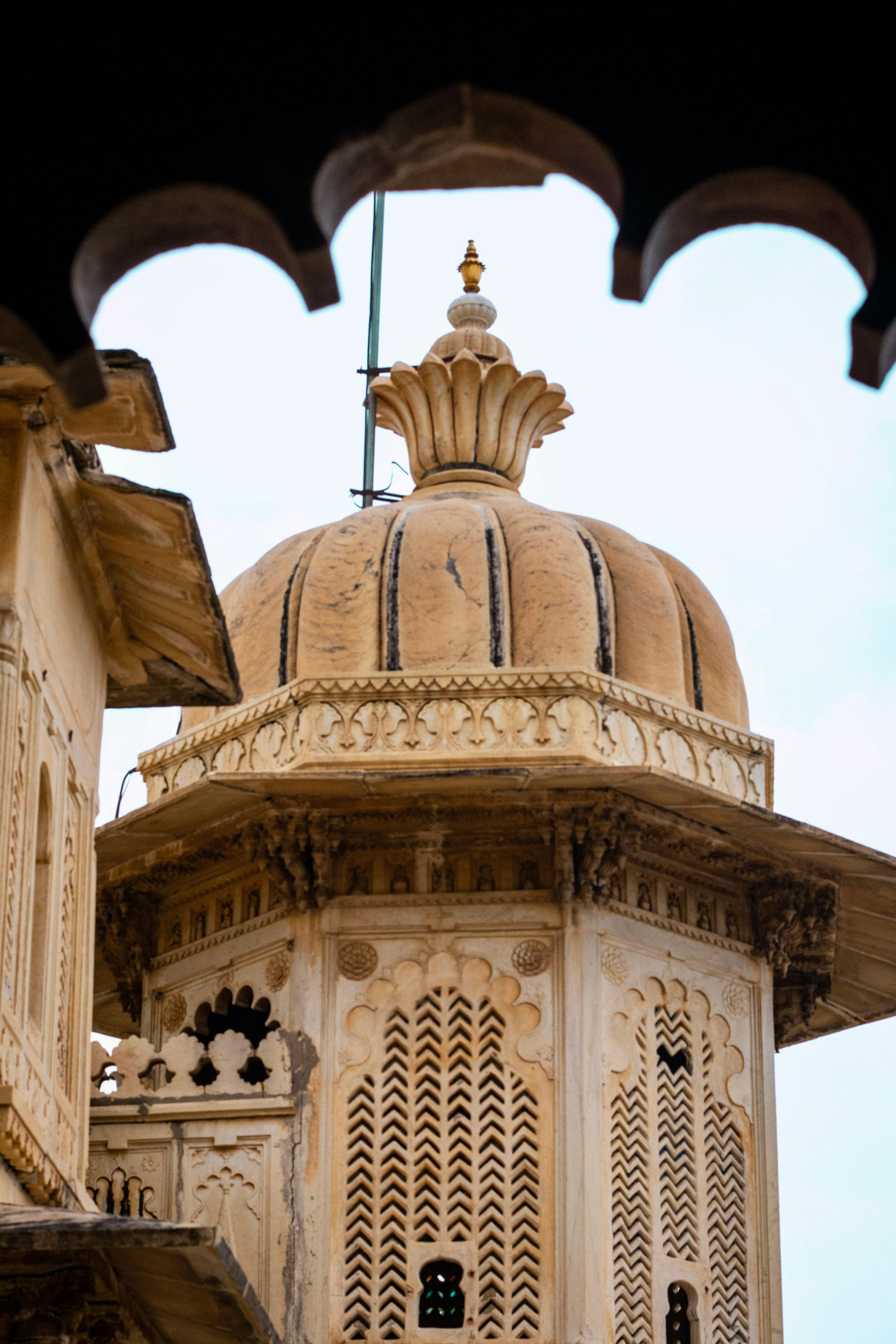 Ornate sandstone dome of a historic Indian building, featuring intricate carvings and arched lattice windows, viewed from below through a decorative archway against a pale sky.