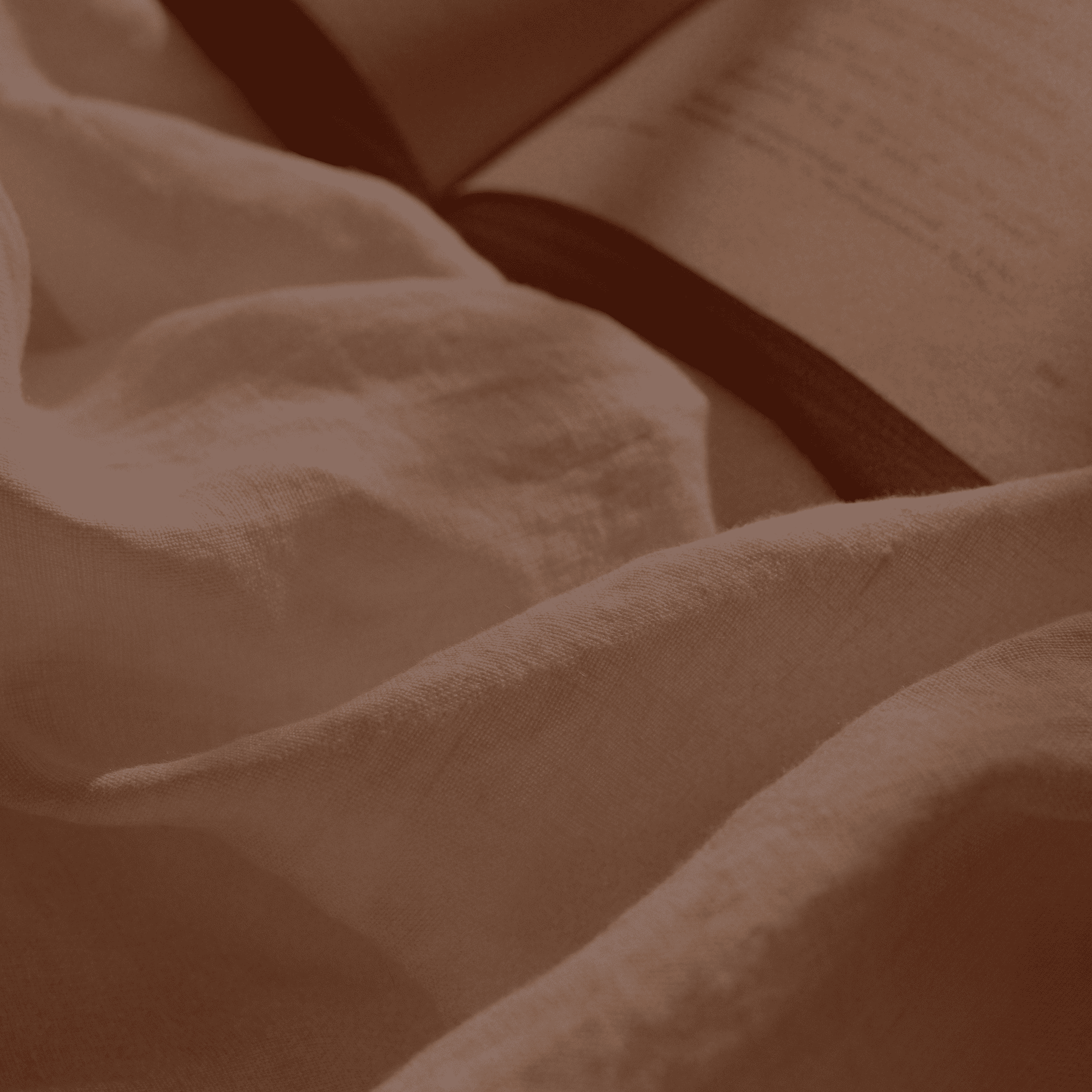 A close-up of draped off-white linen fabric with an open book in the background, softly lit in warm brown tones.