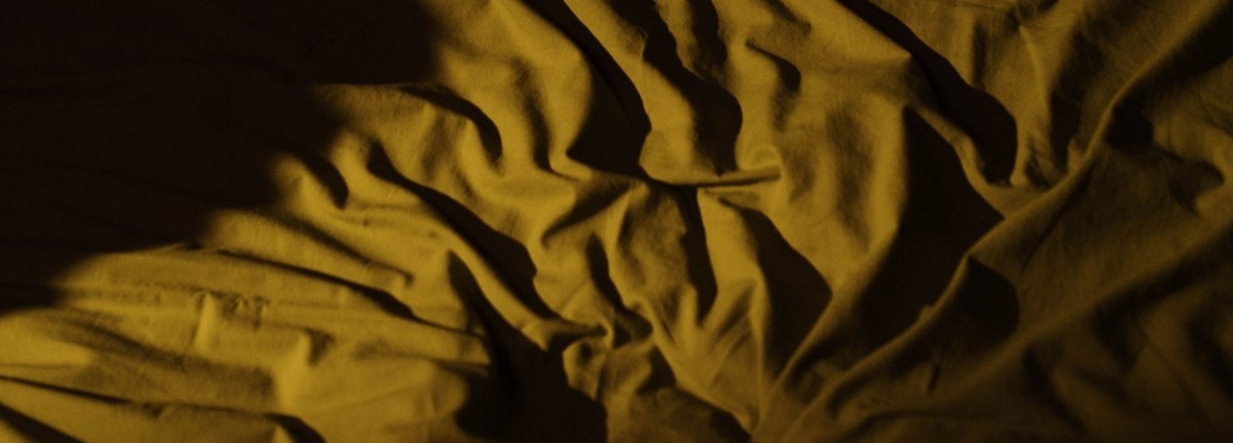 Close-up of rumpled fabric in a mustard yellow or ochre tone under dramatic warm lighting, with deep shadows across the folds.