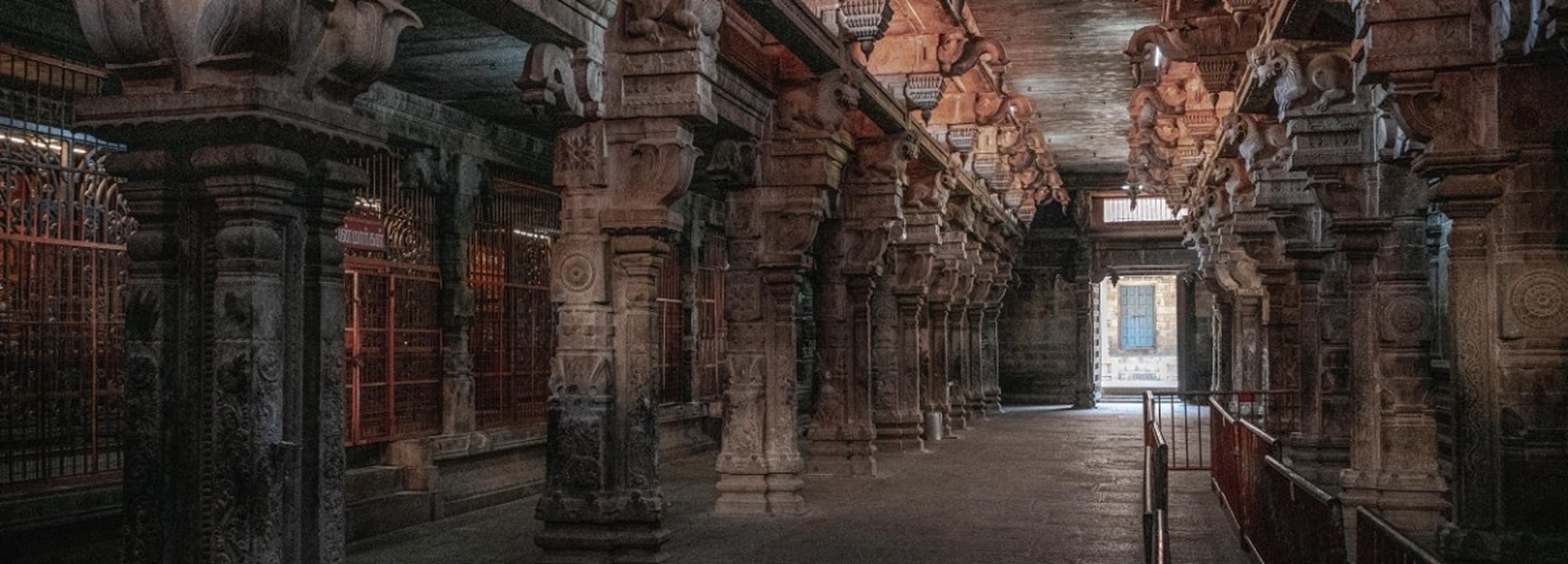 A long stone corridor inside a historic temple or palace, with carved pillars, ornate architectural details, and a bright opening at the far end.