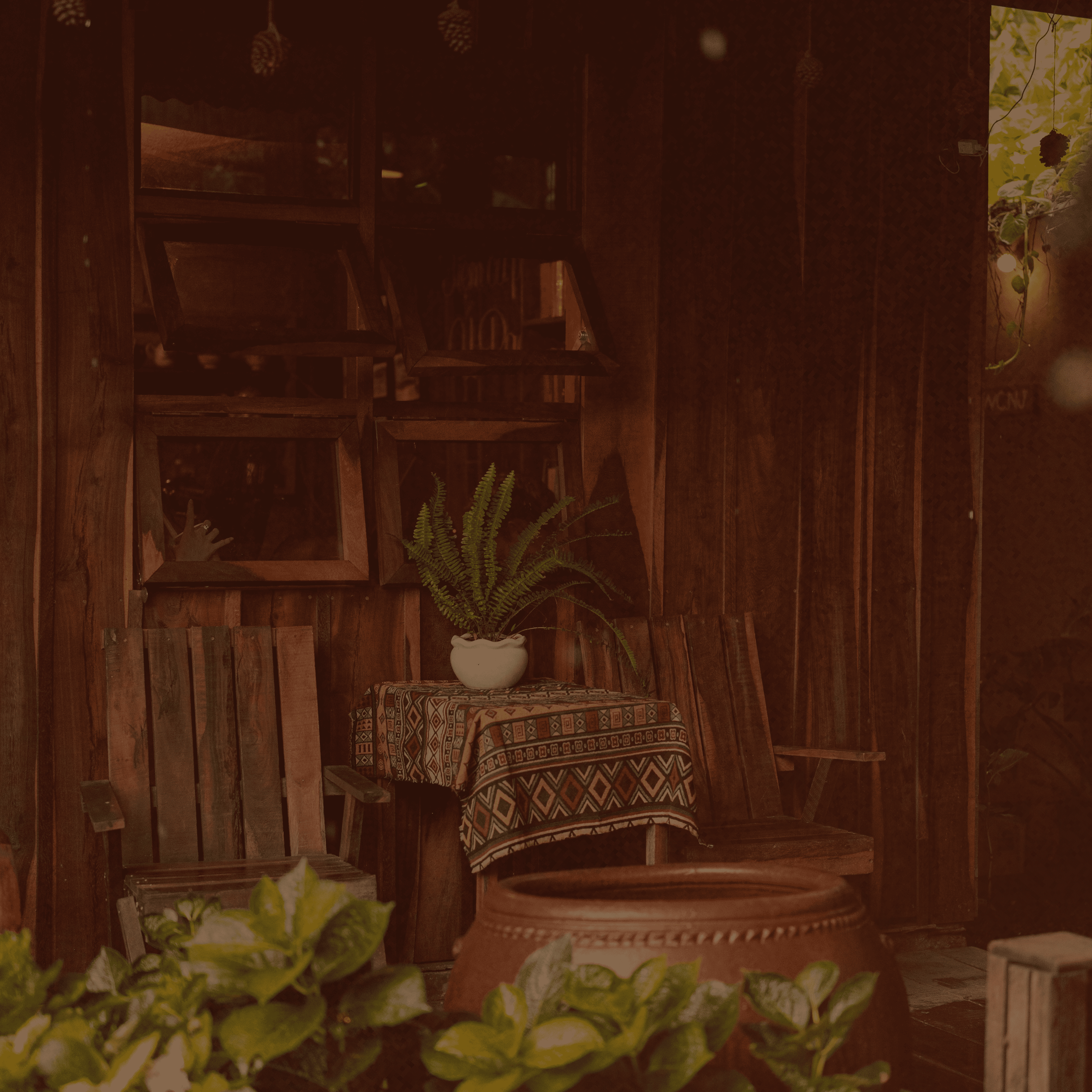 A rustic wooden interior corner with two wooden chairs, a small table covered in a patterned textile, a potted fern in a white pot, and a large terracotta planter in the foreground. The scene is dimly lit with warm ambient light and blurred greenery in the immediate foreground.